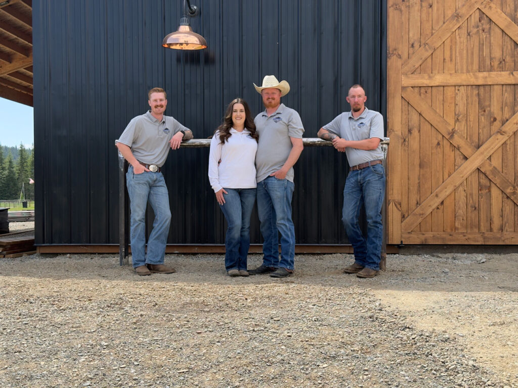 Four members of the Northwest Spray Foam & Insulation team, three men and one woman, pose for a photo in front of a modern barn with black metal siding and large wooden doors. They are dressed in company apparel and casual western wear, standing on a gravel driveway.