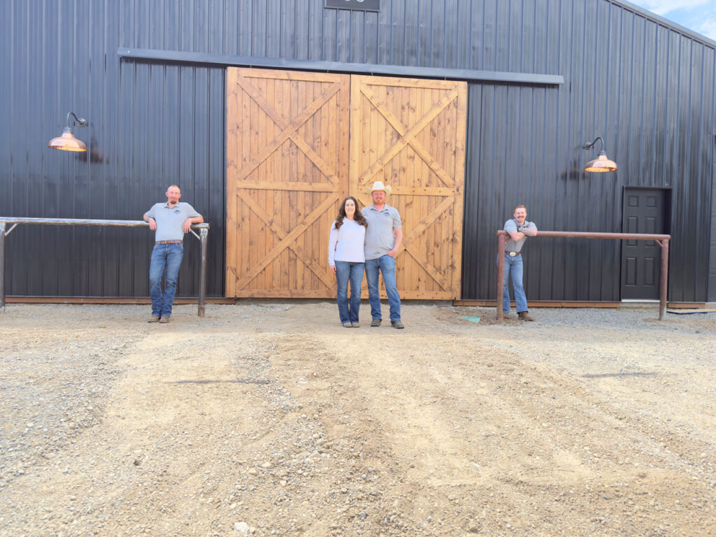 The four-person team from Northwest Spray Foam & Insulation stands in front of a large, dark-colored barn featuring prominent light-wood sliding doors. Two team members stand centrally while the other two lean against railings on either side.