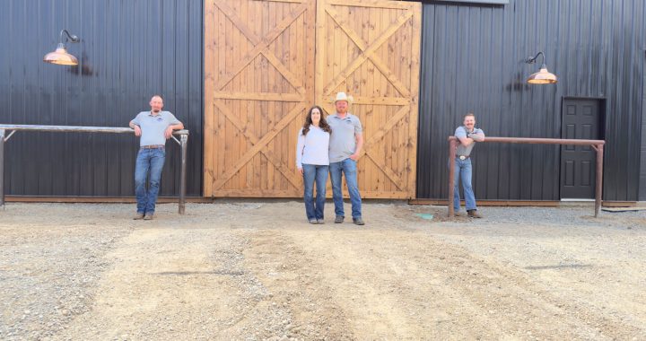 The four-person team from Northwest Spray Foam & Insulation stands in front of a large, dark-colored barn featuring prominent light-wood sliding doors. Two team members stand centrally while the other two lean against railings on either side.