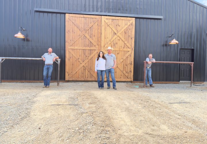 The four-person team from Northwest Spray Foam & Insulation poses proudly in front of a completed project—a modern barn with dark metal siding and large, light-wood sliding doors. They are dressed in company apparel on a gravel driveway.