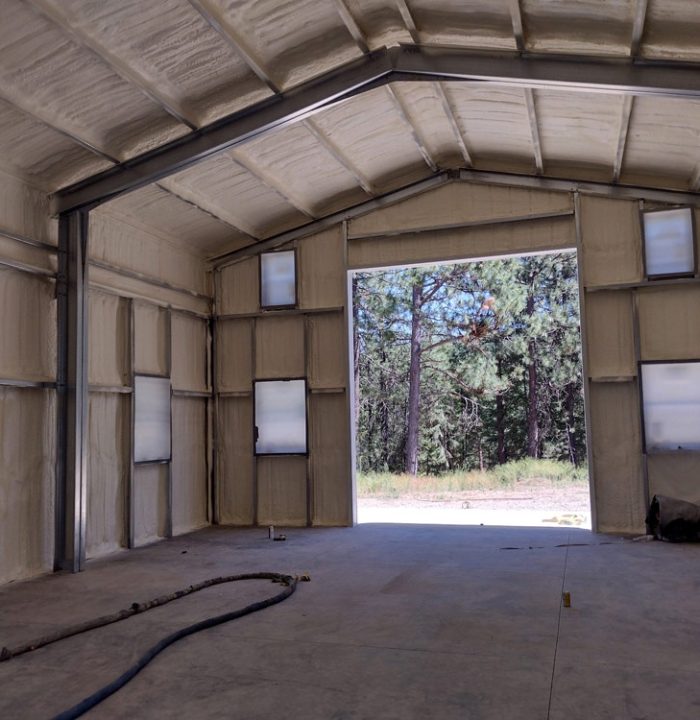 A wide-angle view from inside a large, insulated garage or workshop. The walls and arched ceiling are coated in spray foam, and a large open doorway looks out onto a sunny, forested landscape.