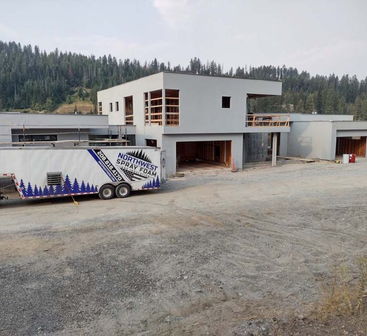 The company's white trailer is parked on a gravel lot in front of a large, modern home under construction. The multi-level house has a stucco exterior and is set against a backdrop of a densely forested hill.