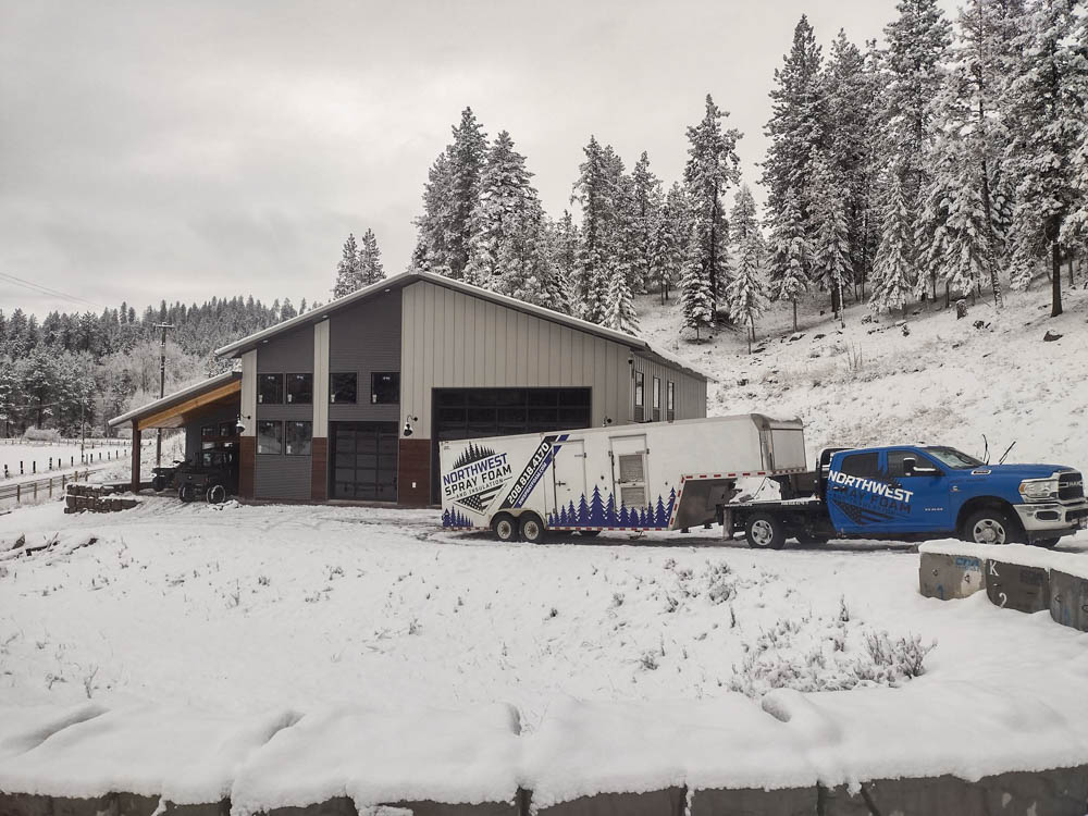 The blue Northwest Spray Foam service truck and branded trailer are parked in the snow outside a modern barndominium-style building. The surrounding landscape, including the pine trees on the hillside, is covered in a fresh layer of snow.