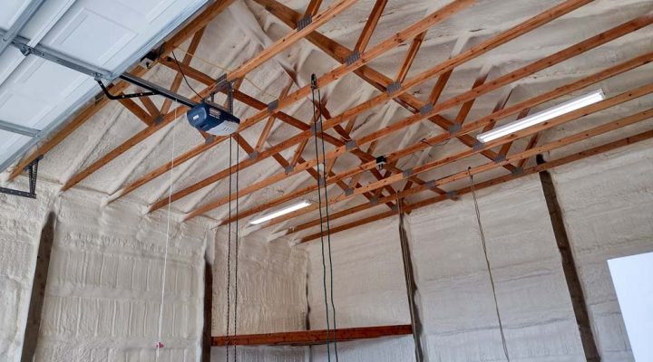 The interior of a garage after being insulated with spray foam. The foam covers the walls and the underside of the roof deck, neatly applied around the exposed wooden trusses. A garage door opener and fluorescent lights are mounted to the ceiling structure.