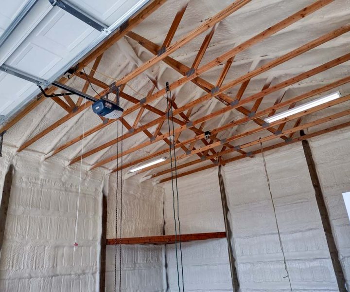 The interior of a garage after being insulated with spray foam. The foam covers the walls and the underside of the roof deck, neatly applied around the exposed wooden trusses. A garage door opener and fluorescent lights are mounted to the ceiling structure.