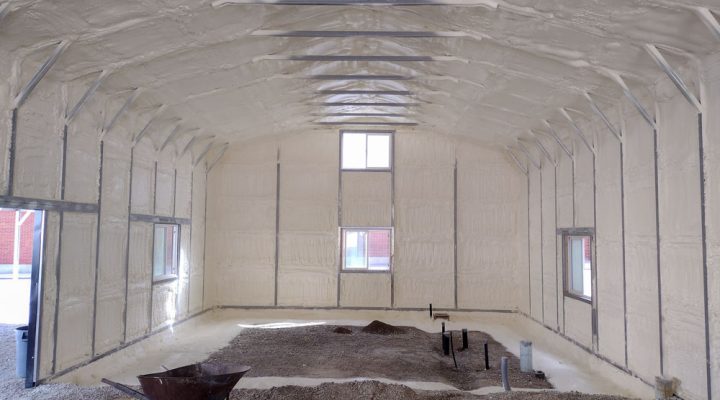 The inside of an arched, Quonset hut-style building after the walls and ceiling have been insulated with spray foam. A wheelbarrow and piles of gravel are on the floor, indicating that the space is still under construction but now has a complete thermal envelope.