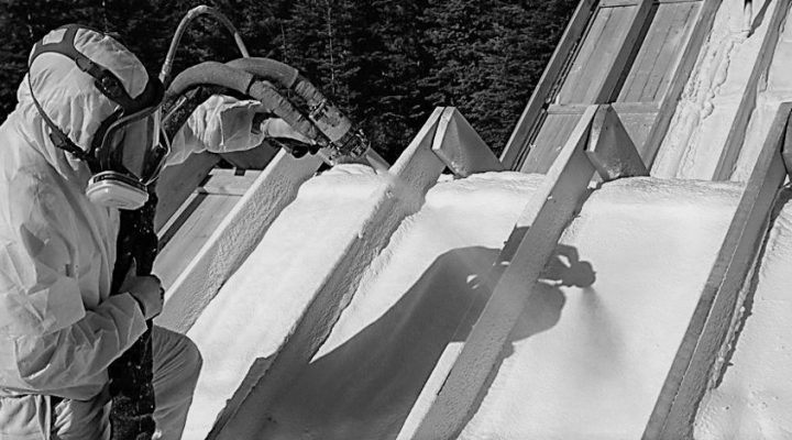 A black and white photo shows a technician in full protective gear, including a respirator, applying spray foam insulation between the rafters of a roof. The image highlights the precision and safety measures involved in the application process.