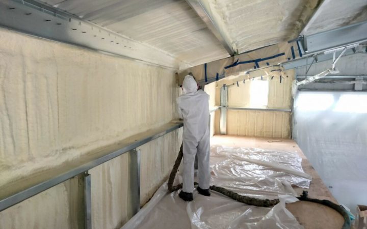 A technician in a protective suit applies spray foam insulation to the upper walls and ceiling of a metal building. The floor is covered with protective plastic sheeting, and windows are masked off to ensure a clean application.