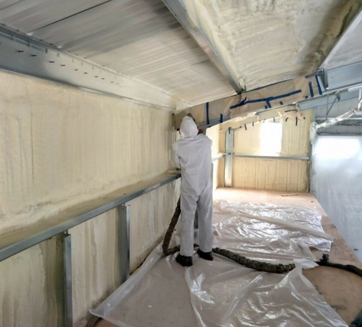A technician in a protective suit applies spray foam insulation to the upper walls and ceiling of a metal building. The floor is covered with protective plastic sheeting, and windows are masked off to ensure a clean application.