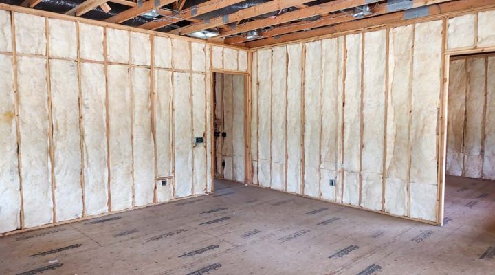 A wide shot of a large, empty room where all the walls have been insulated with spray foam applied between the wooden studs. The exposed ceiling trusses and AdvanTech subfloor indicate the project is ready for the next phase of construction.