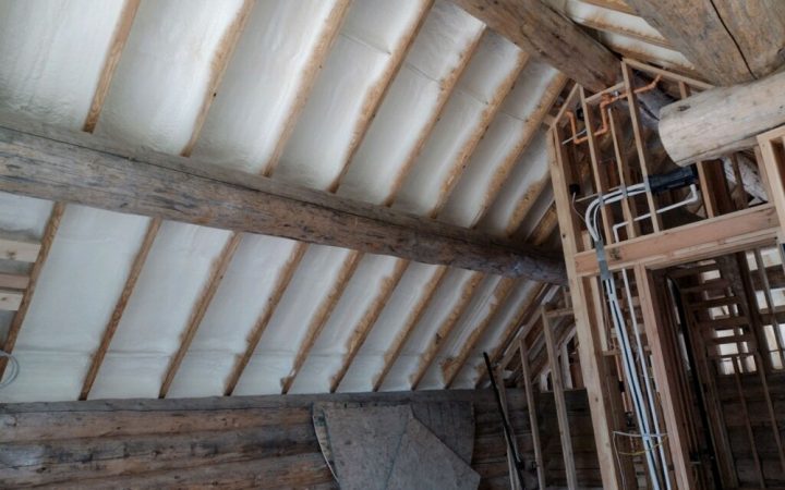 An interior view of an attic space in a log home after insulation. Spray foam fills the cavities between the wooden rafters of the sloped ceiling, while large, rustic log beams remain exposed. New framing for interior rooms is visible on the right.