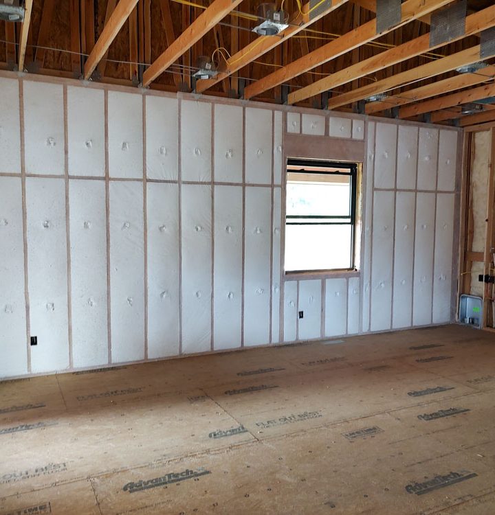An unfinished room displays walls insulated with spray foam and covered with rigid foam boards. The floor is made of AdvanTech subflooring, and plumbing pipes are visible in one corner, indicating preparation for interior finishing.