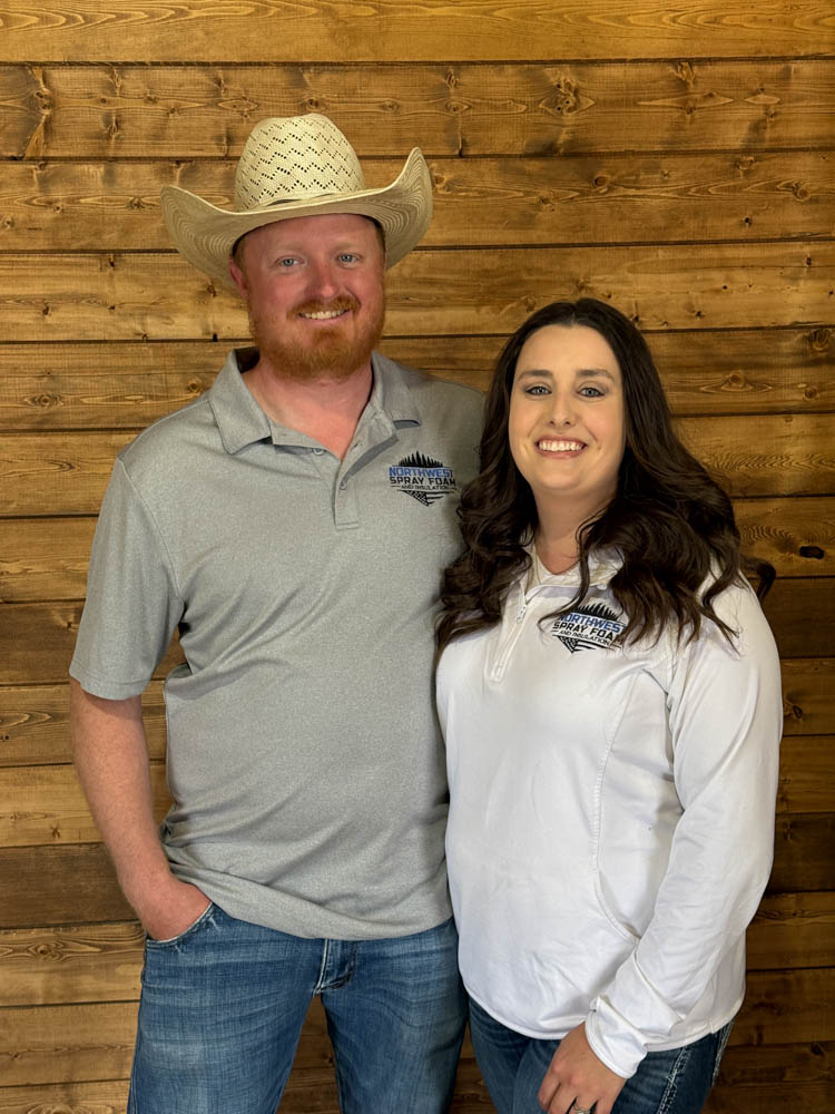 A smiling man and woman, presumably the founders or owners of Northwest Spray Foam & Insulation, pose together against a warm-toned wood-paneled wall. Both are wearing company-branded polo shirts, with the man also sporting a cowboy hat.