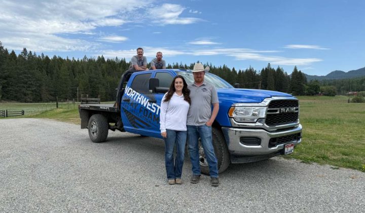 The full team of four from Northwest Spray Foam & Insulation poses with their branded blue Ram truck. Two members stand in front of the vehicle while the other two are in the truck bed, set against a backdrop of green fields and forests under a partly cloudy sky.