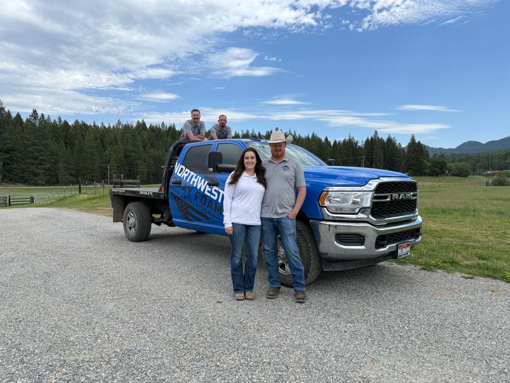 The full team of four from Northwest Spray Foam & Insulation poses with their branded blue Ram truck. Two members stand in front of the vehicle while the other two are in the truck bed, set against a backdrop of green fields and forests under a partly cloudy sky.